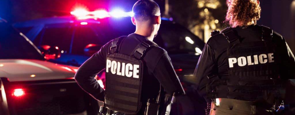 Rear view of two multiracial police officers standing side by side in front of their police vehicles with emergency lights on. They are working at night in a city. The policewoman is mixed race, African-American, Asian and Hispanic, in her 40s. Her partner is a young Hispanic man in his 20s.